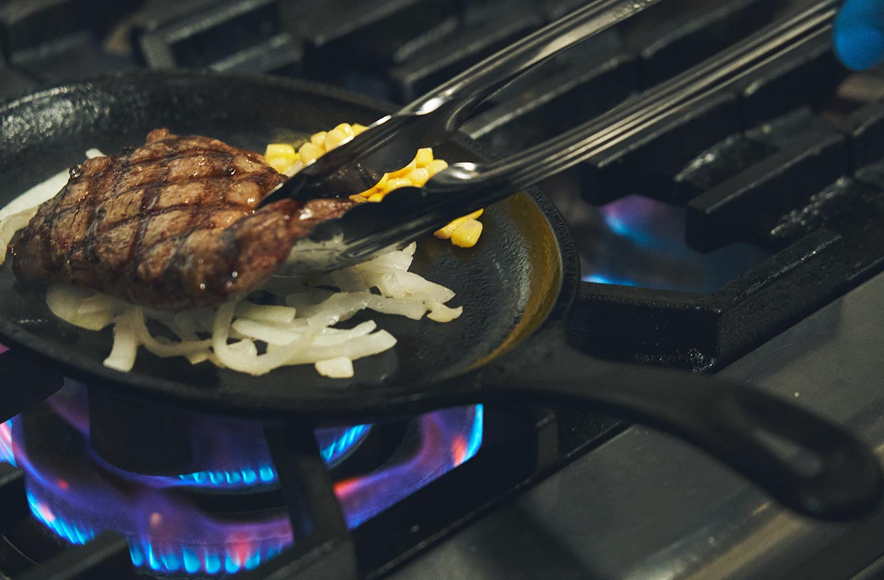 ステーキは、グリル板で焼いた後、タマネギとコーンをのせたアツアツの鉄板にのせて提供する。